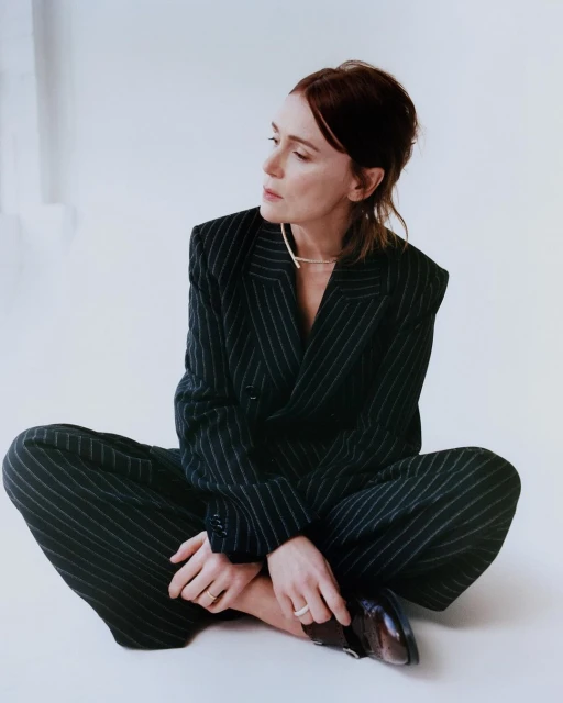 Stylish woman in a pinstripe suit sits cross-legged on a white surface, looking off to the side, showcasing fashion and modern professional attire.