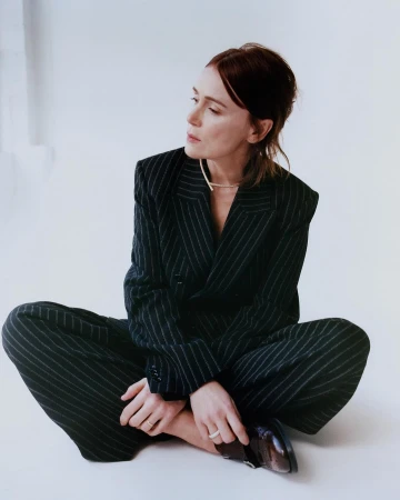 Stylish woman in a pinstripe suit sits cross-legged on a white surface, looking off to the side, showcasing fashion and modern professional attire.