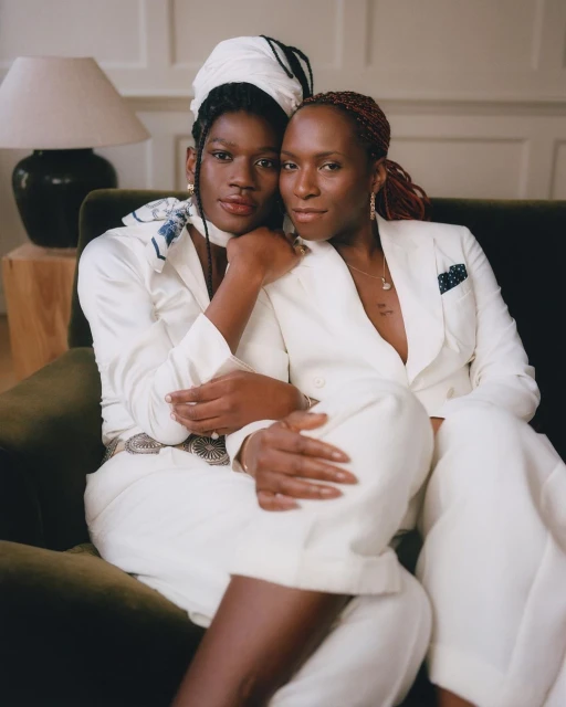 Two African-descent women in suits pose on a green couch indoors, bathed in soft window light. One wears a headscarf and patterned scarf.