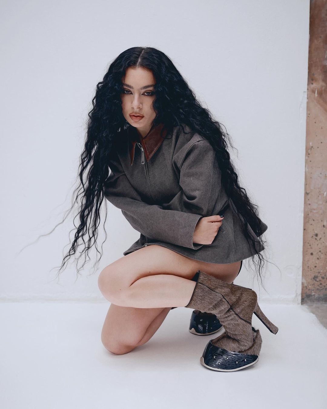 Woman with long dark wavy hair in a grey jacket and platform boots crouching indoors against a neutral wall in a fashion shot.