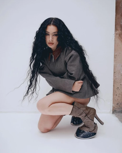 Woman with long dark wavy hair in a grey jacket and platform boots crouching indoors against a neutral wall in a fashion shot.
