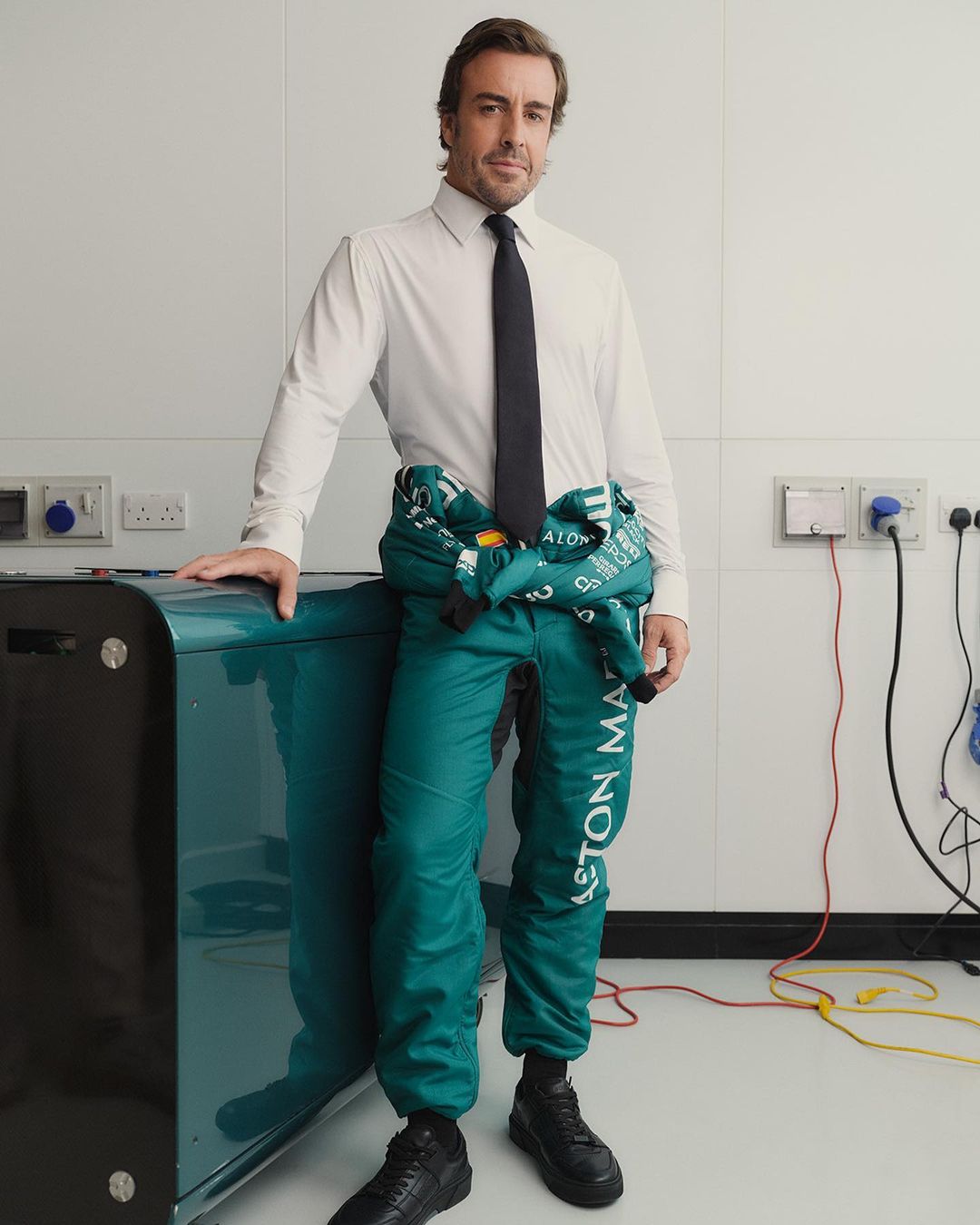 Professional portrait of a male in a semi-formal outfit with a racing jumpsuit over his shoulder, indoors, posing near a teal-colored object.