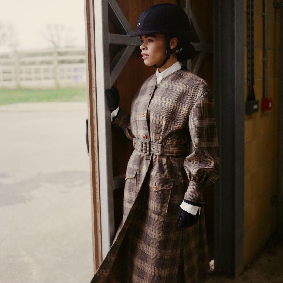 Woman in equestrian helmet and plaid coat stands in barn doorway, looking out; neutral tones, diffused natural light, medium shot.
