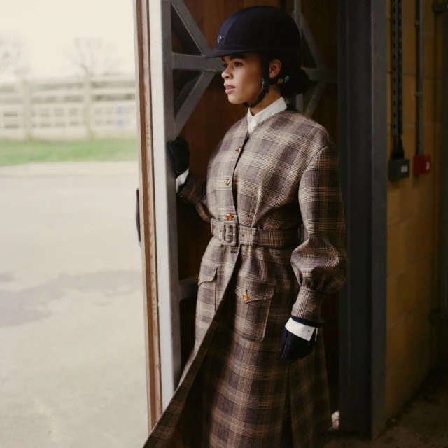 Woman in equestrian helmet and plaid coat stands in barn doorway, looking out; neutral tones, diffused natural light, medium shot.