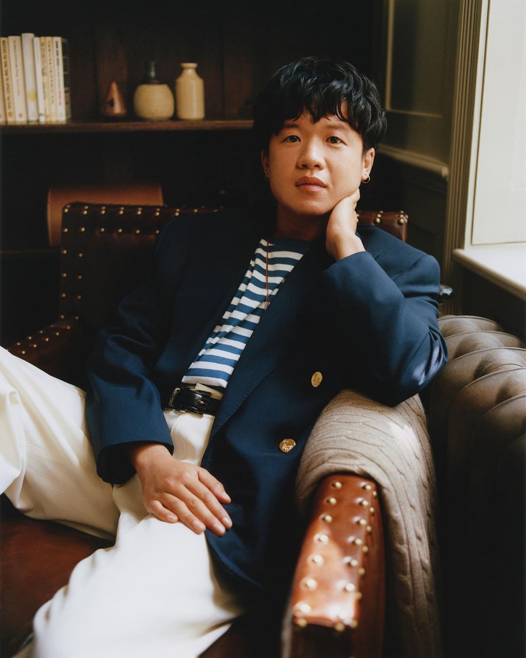 Portrait of an adult person with short dark hair, wearing a navy blazer and white pants, sitting on a leather studded chair.