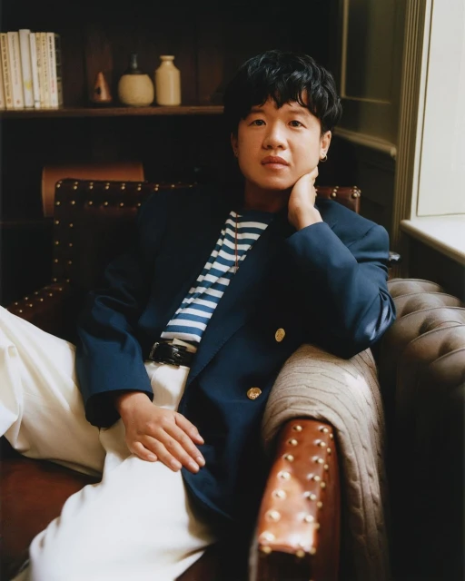 Portrait of an adult person with short dark hair, wearing a navy blazer and white pants, sitting on a leather studded chair.