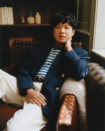Portrait of an East Asian man in his 30s seated in a leather armchair wearing a navy blazer and striped t-shirt