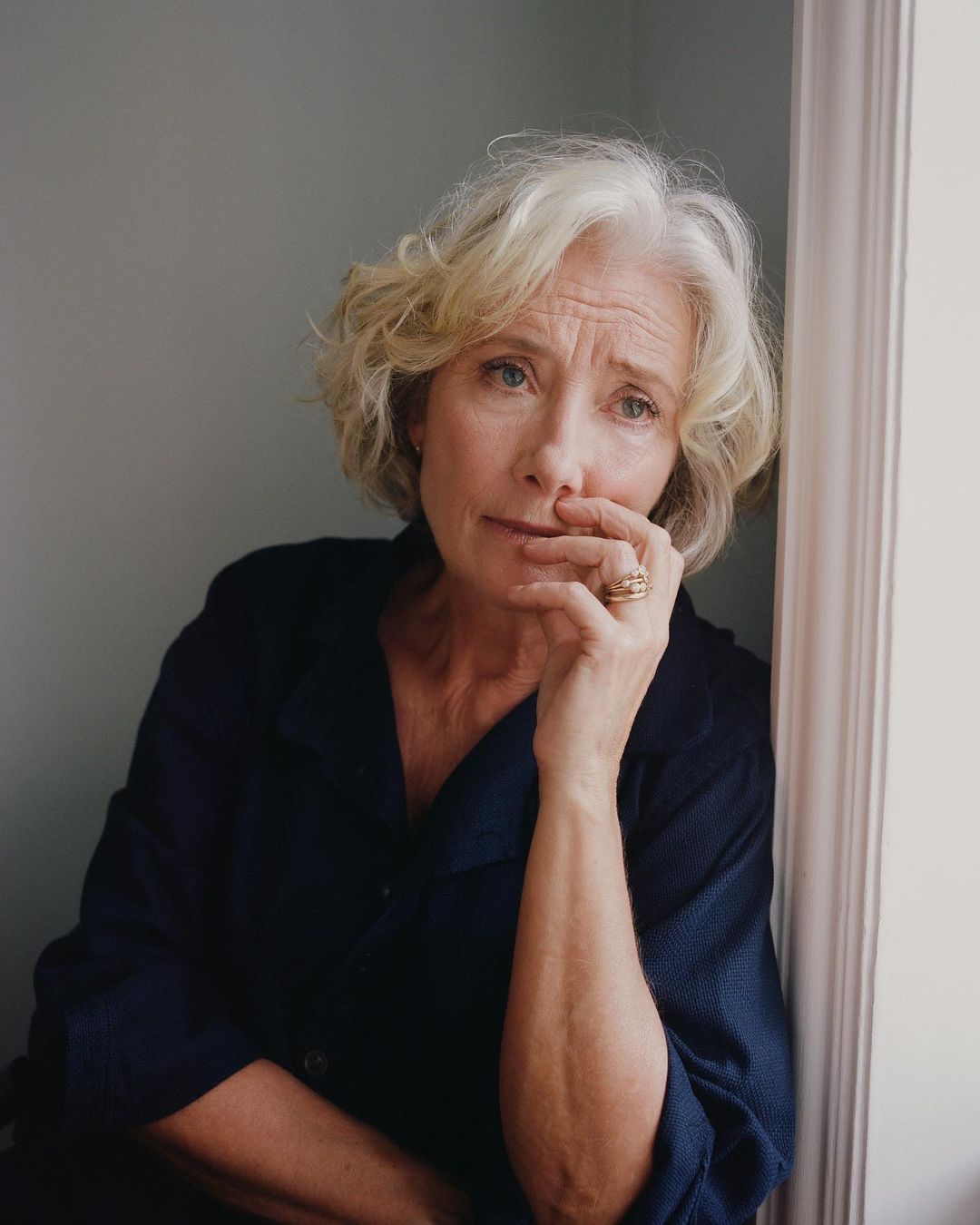 Emma Thompson portrait in a dark blue shirt with curly grey hair, leaning against a white wall, looking thoughtfully at the camera.