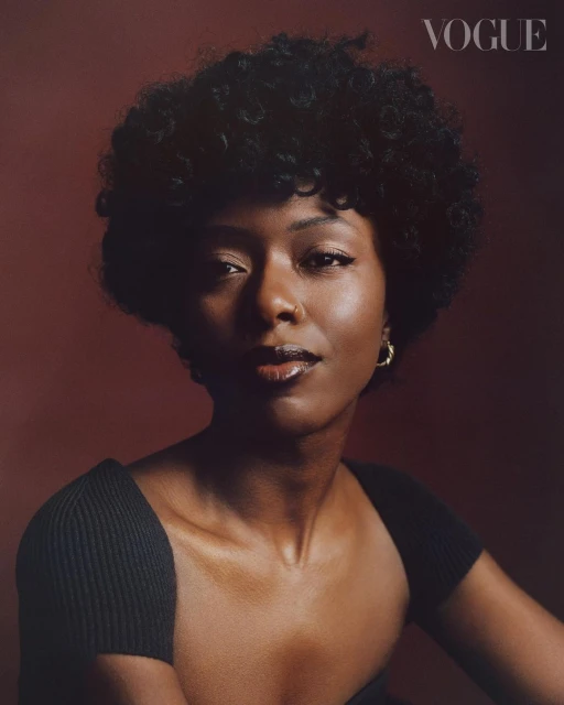 Headshot of a young African American woman with dark curly hair, gold earrings, wearing a black top against a maroon background.
