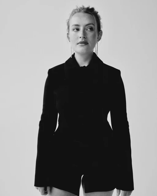 Monochrome portrait of a woman in a black blazer and shorts against a stark white backdrop, with her gaze directed off-camera.