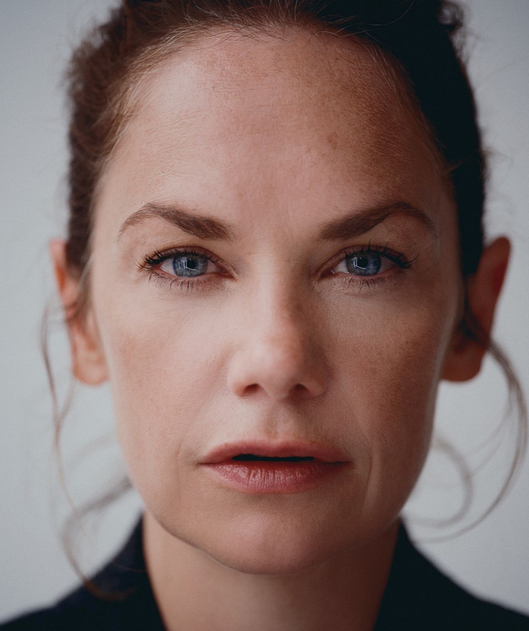 Close-up portrait of a fair-skinned adult female with red hair pulled back, featuring blue eyes and a blazer against a neutral backdrop.