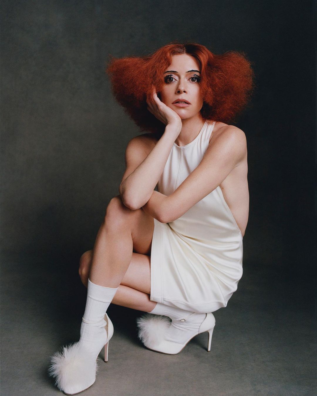 Woman with red hair poses in a short white dress and fuzzy platform heels against a neutral background in a studio portrait.