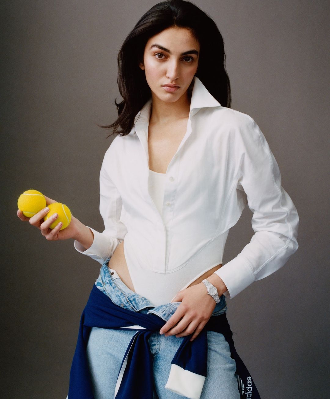 A woman in a white bodysuit and shirt holds two tennis balls against a neutral backdrop, styled with light-wash jeans and an Adidas jacket.