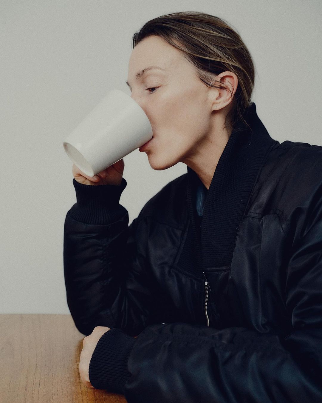 Woman wearing a black bomber jacket drinking from a white mug at a wooden table against a plain wall.
