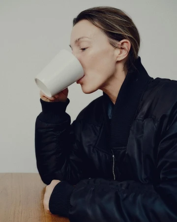 Woman wearing a black bomber jacket drinking from a white mug at a wooden table against a plain wall.