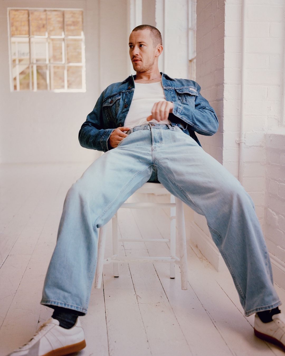 Man sitting on a stool in jeans and a denim jacket in a white room with large windows. Casual fashion in a minimalist space.