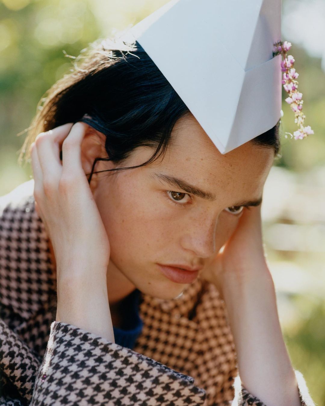 Young adult with a paper hat decorated with pink flowers poses outdoors wearing a houndstooth jacket in medium close-up.