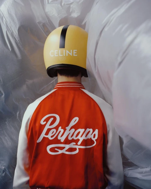 Person wearing a yellow helmet with 'CELINE' text and a red and white varsity jacket with 'Perhaps' text in a studio setting.