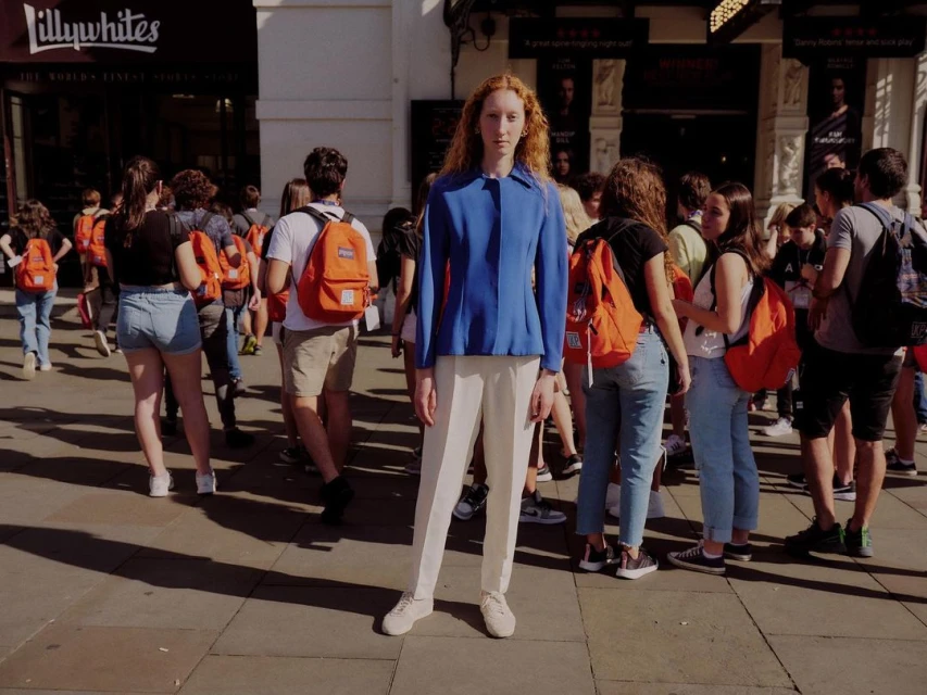 Red-haired person in a blue jacket and white trousers stands surrounded by a crowd on a city street with shopfronts behind.