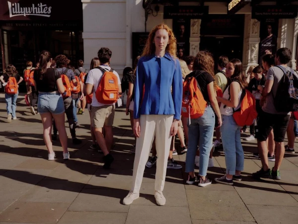 Red-haired person in a blue jacket and white trousers stands surrounded by a crowd on a city street with shopfronts behind.