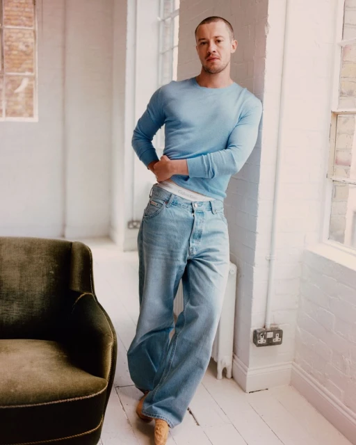Man in casual light blue shirt and jeans poses in a light-filled room with chair near a window and white brick wall. 