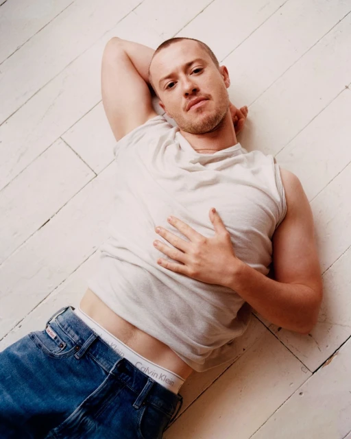 Man lying on a white wooden floor, wearing a white sleeveless shirt and blue jeans, with a hand on his stomach and head resting.