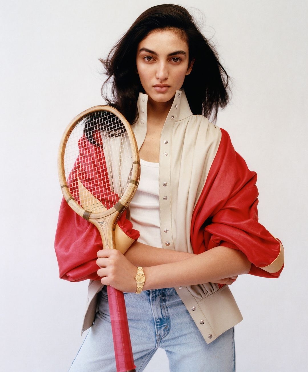Young woman poses in a studio wearing a red and white jacket, light jeans, and holding a tennis racket in a fashion shot.