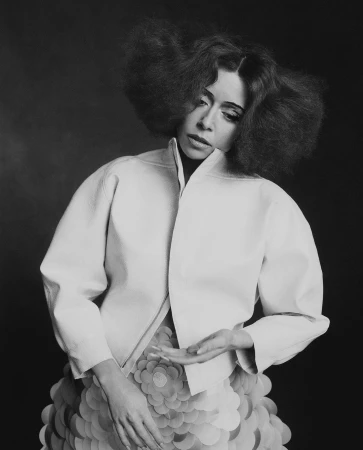Portrait of a woman with voluminous dark hair wearing a light-colored high-collar jacket against a dark background.