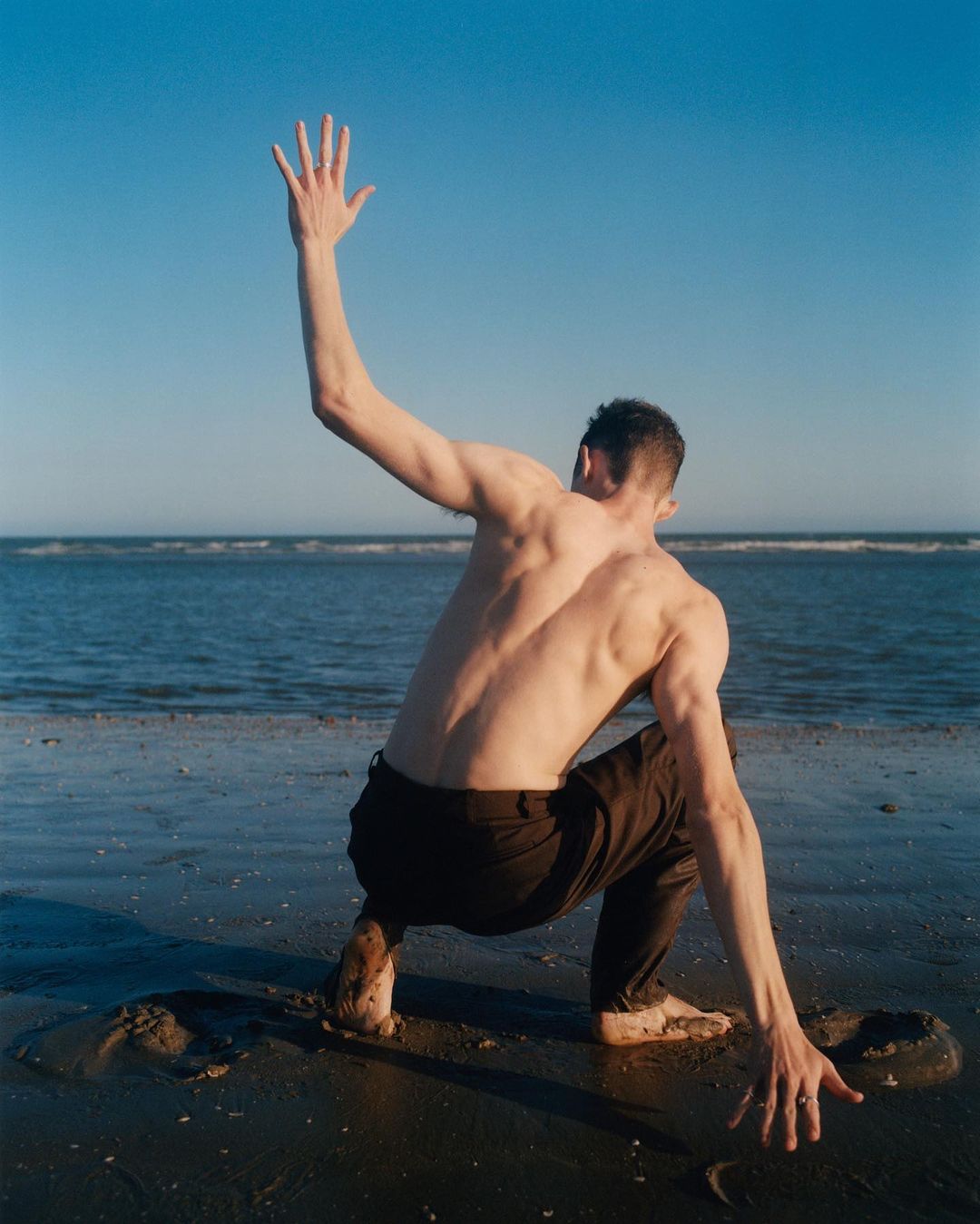 Man with bare torso on a sandy beach, in a crouched position with one arm raised, facing the ocean under a clear blue sky.