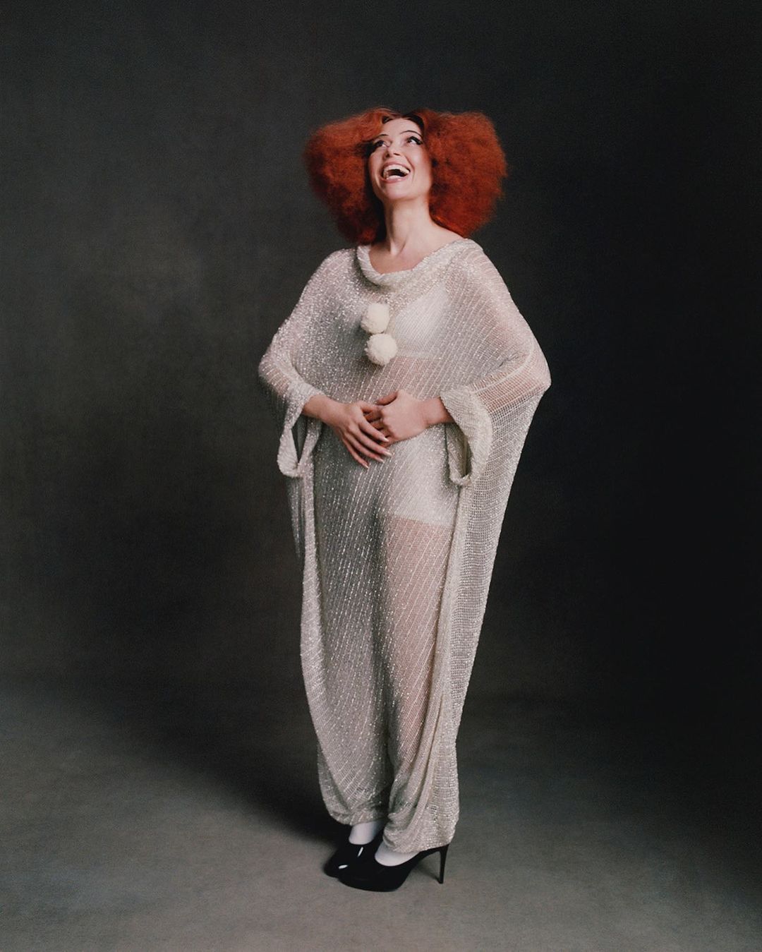 Full shot of an adult woman with red hair wearing a translucent white dress, smiling and looking up against a dark backdrop.