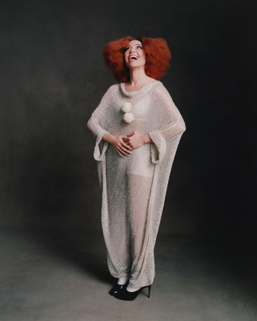 Full shot of an adult woman with red hair wearing a translucent white dress, smiling and looking up against a dark backdrop.
