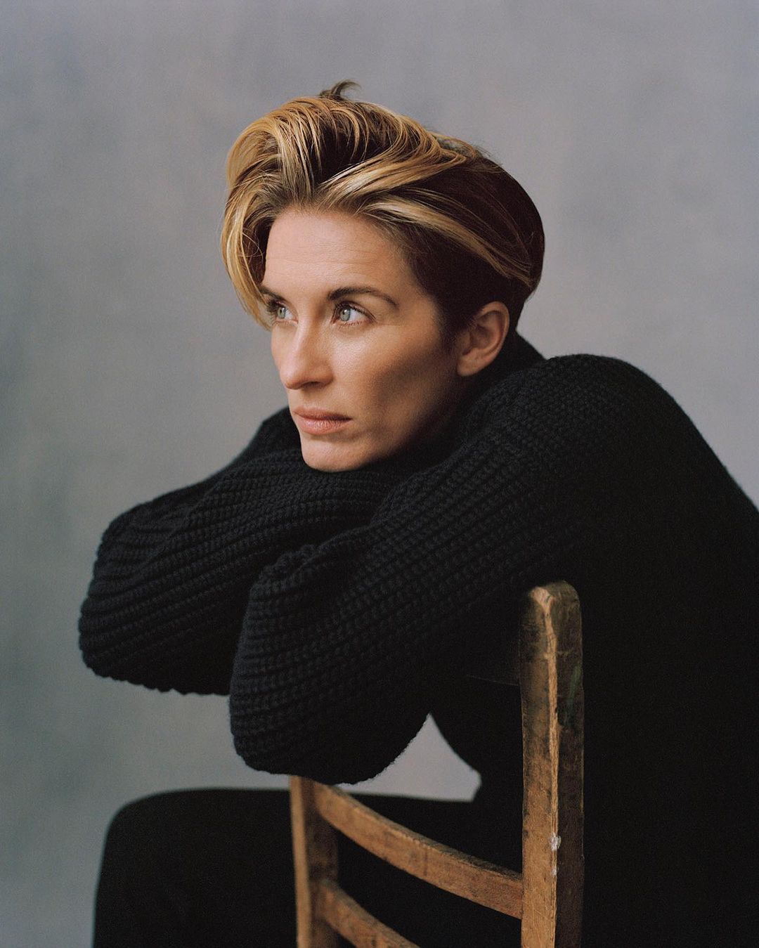 Portrait of a blonde woman with a contemplative expression, wearing a black knit sweater and leaning on a wooden chair against a grey backdrop.