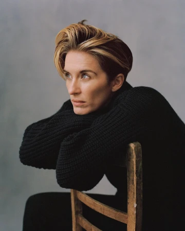 Portrait of a blonde woman with a contemplative expression, wearing a black knit sweater and leaning on a wooden chair against a grey backdrop.
