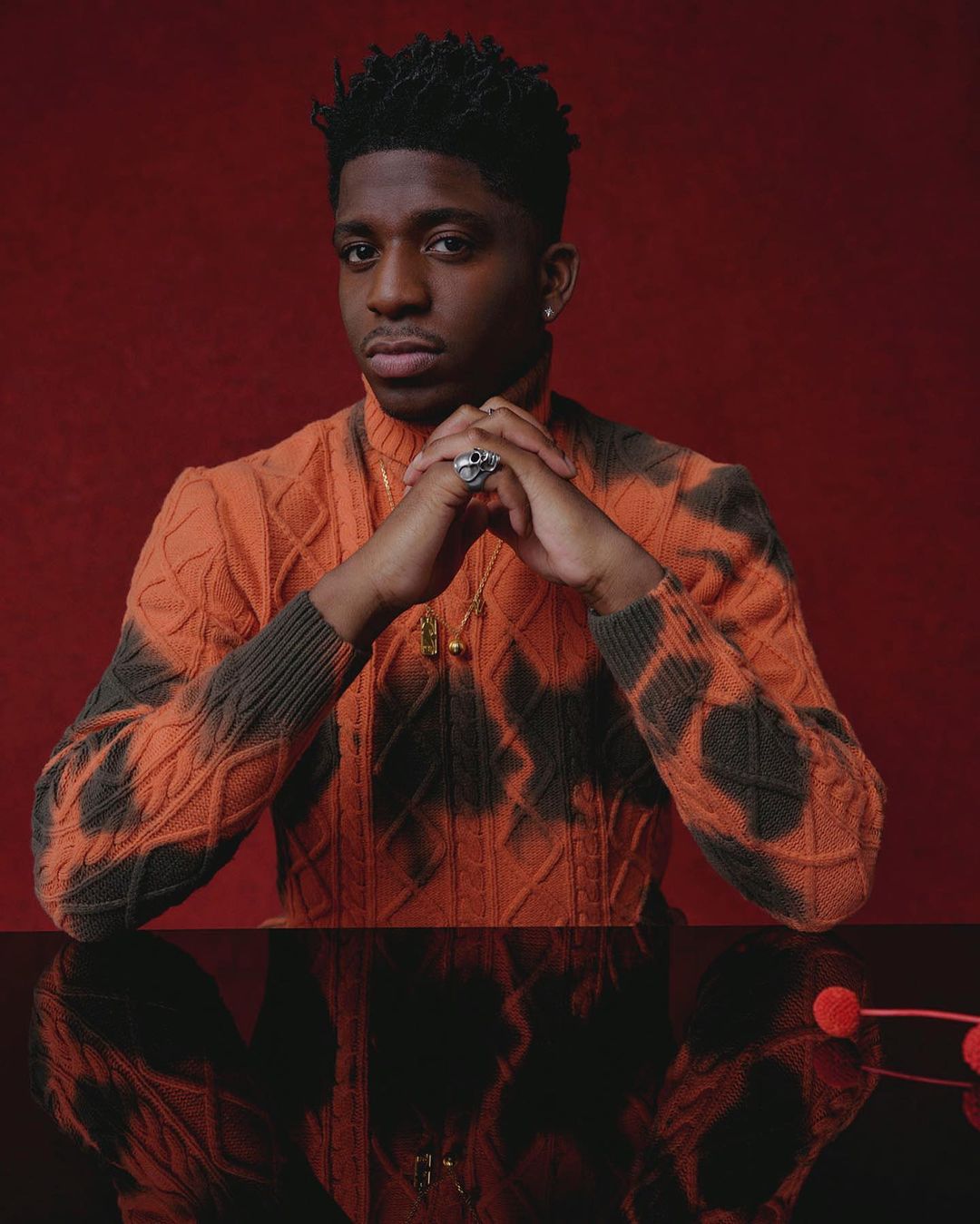 Portrait of a Black man with short dark hair wearing an orange patterned turtleneck sweater, with his hands clasped.