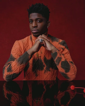 Portrait of a Black man with short dark hair wearing an orange patterned turtleneck sweater, with his hands clasped.
