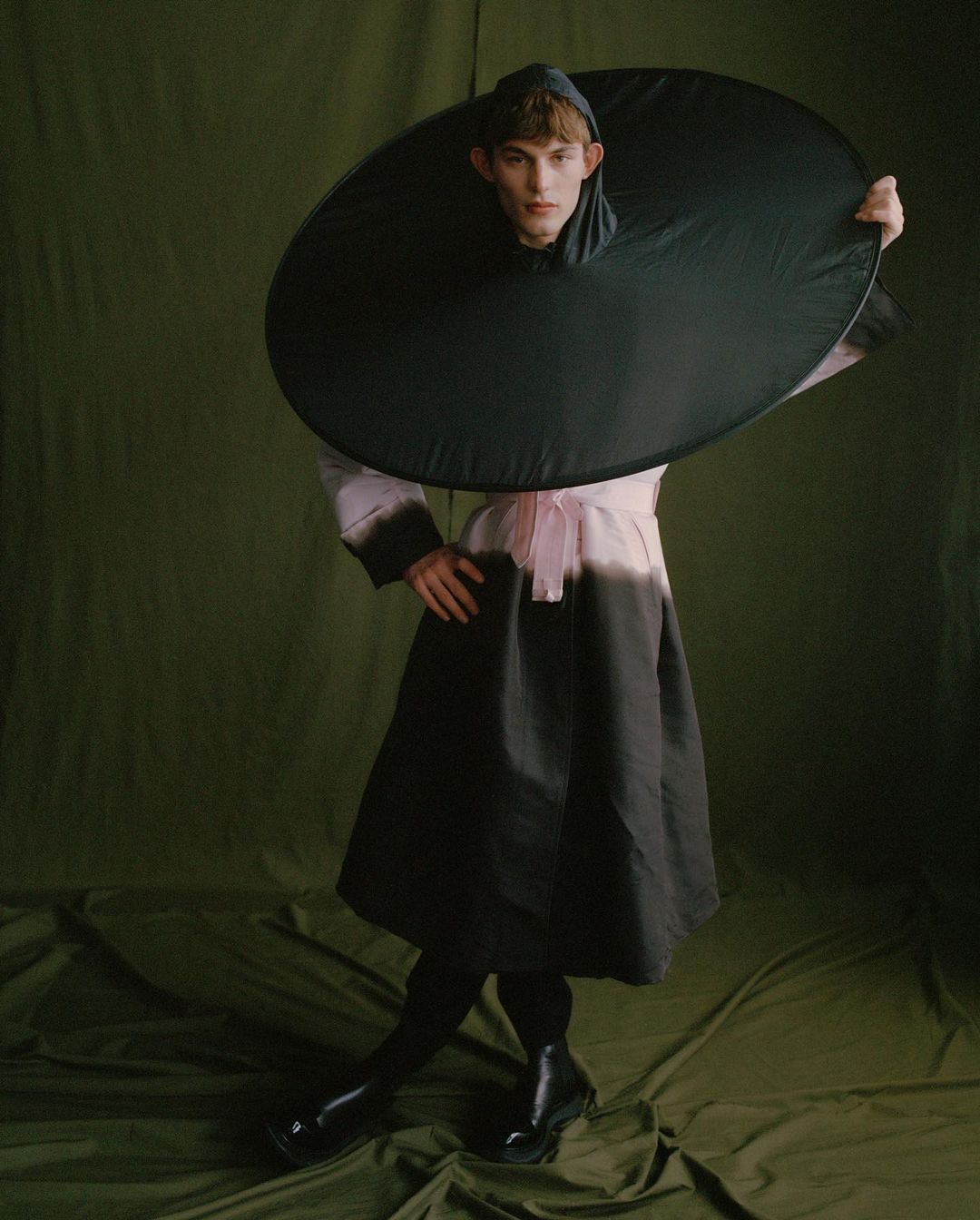 Fashionable young adult in a striking black-and-pink dress stands against an olive-green backdrop, enhanced by studio lighting and unique styling.