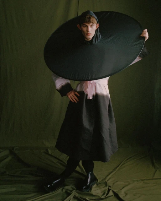 Fashionable young adult in a striking black-and-pink dress stands against an olive-green backdrop, enhanced by studio lighting and unique styling.