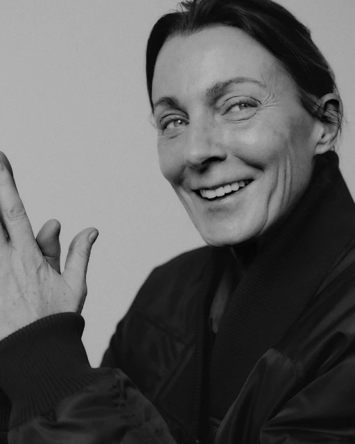 Portrait of a smiling mature adult woman in a black bomber jacket, with a neutral background, captured in a black and white photograph.