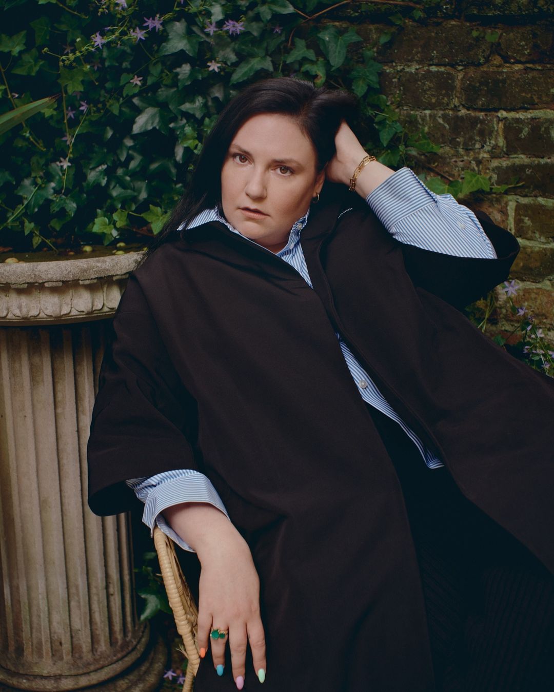 Portrait of a woman sitting outdoors against a leafy brick wall backdrop, wearing a dark coat over a striped shirt, accessorized with a ring.