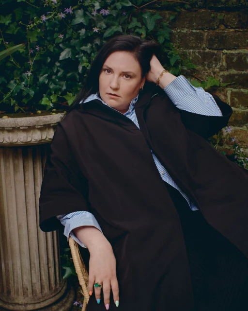 Portrait of a woman sitting outdoors against a leafy brick wall backdrop, wearing a dark coat over a striped shirt, accessorized with a ring.