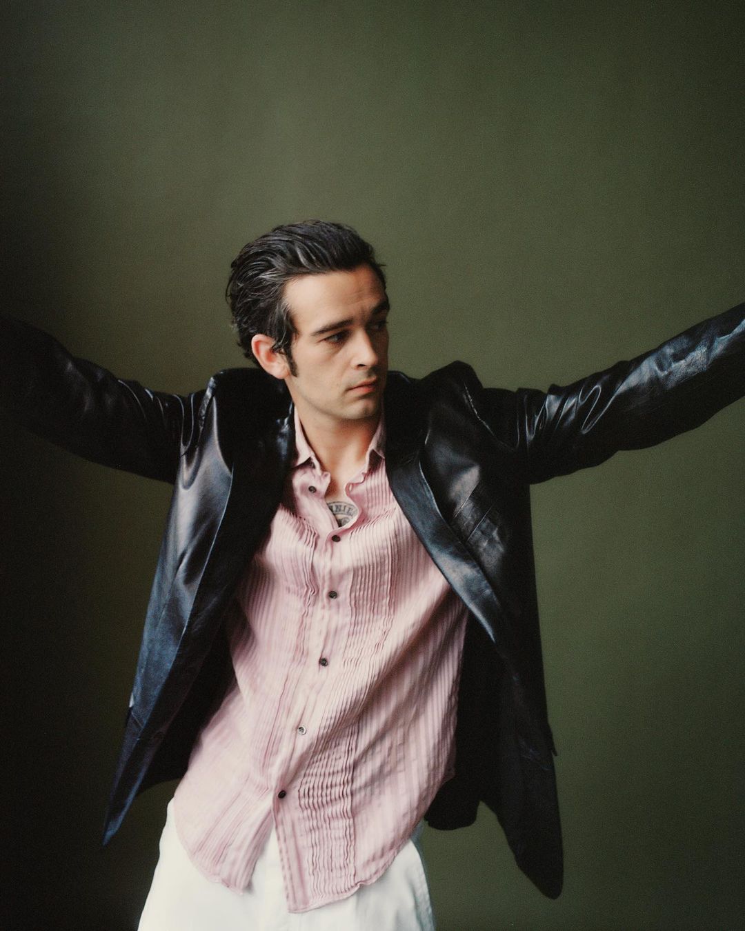 Portrait of Matty Healy wearing a black leather jacket and pink shirt with arms outstretched.