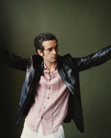 Portrait of Matty Healy wearing a black leather jacket and pink shirt with arms outstretched.