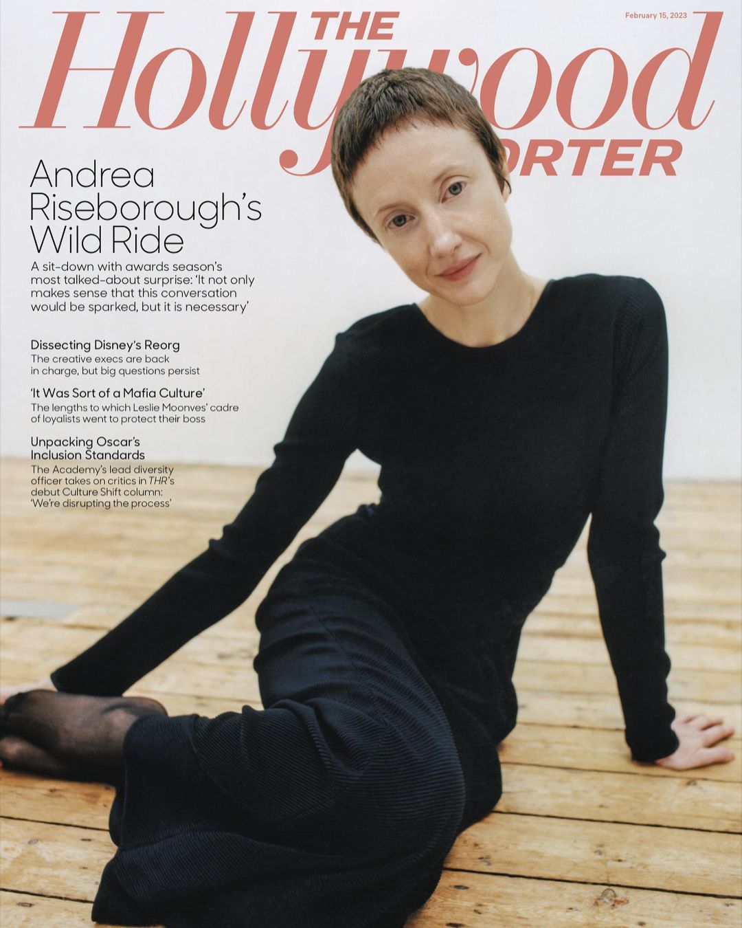 Portrait of Andrea Riseborough seated on a wooden floor against a white wall wearing a long sleeve black dress.