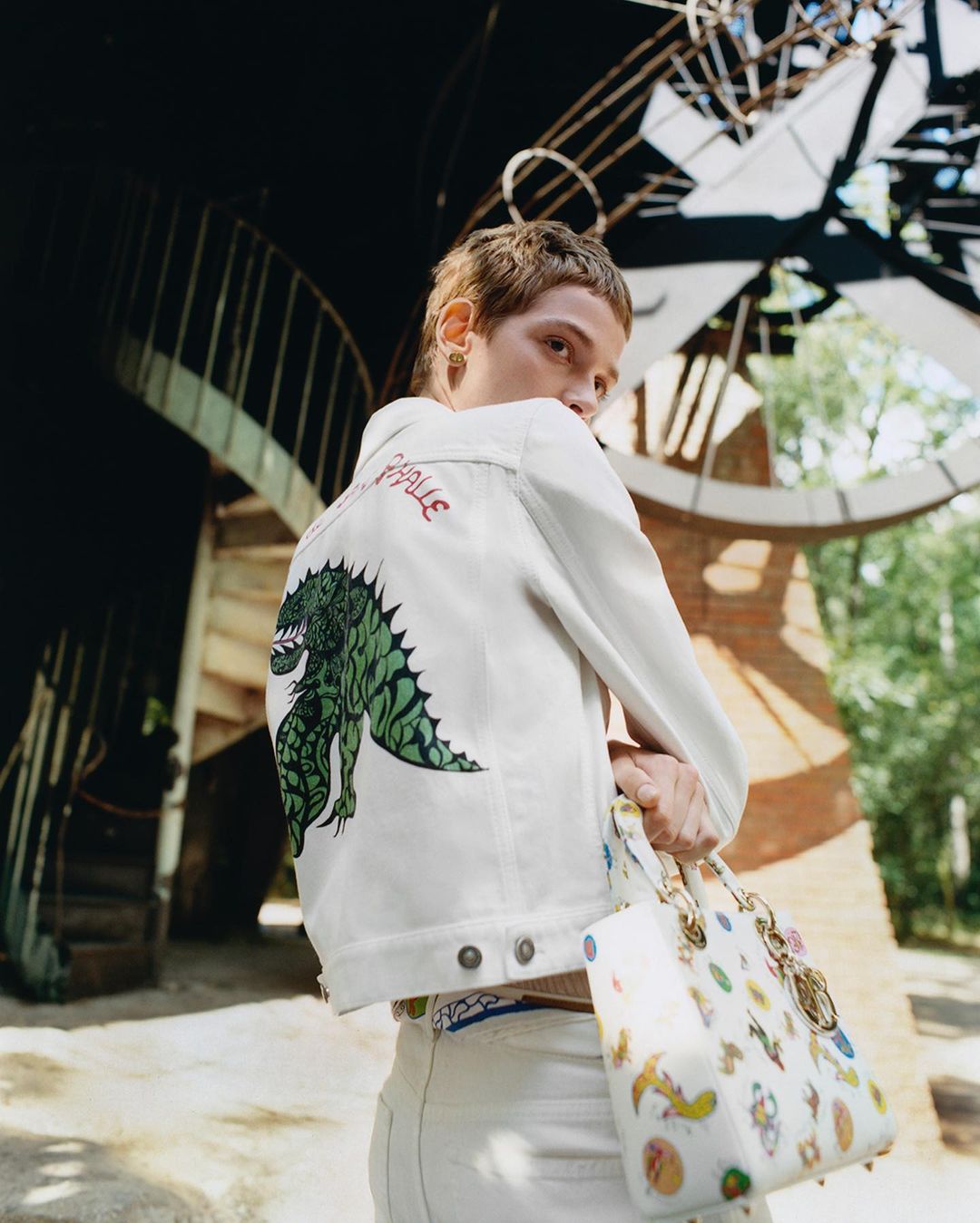 Young adult posing, wearing a white jacket with a dragon design, holding a decorated handbag, outdoors, with a light-toned background.
