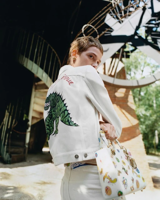 Young adult posing, wearing a white jacket with a dragon design, holding a decorated handbag, outdoors, with a light-toned background.