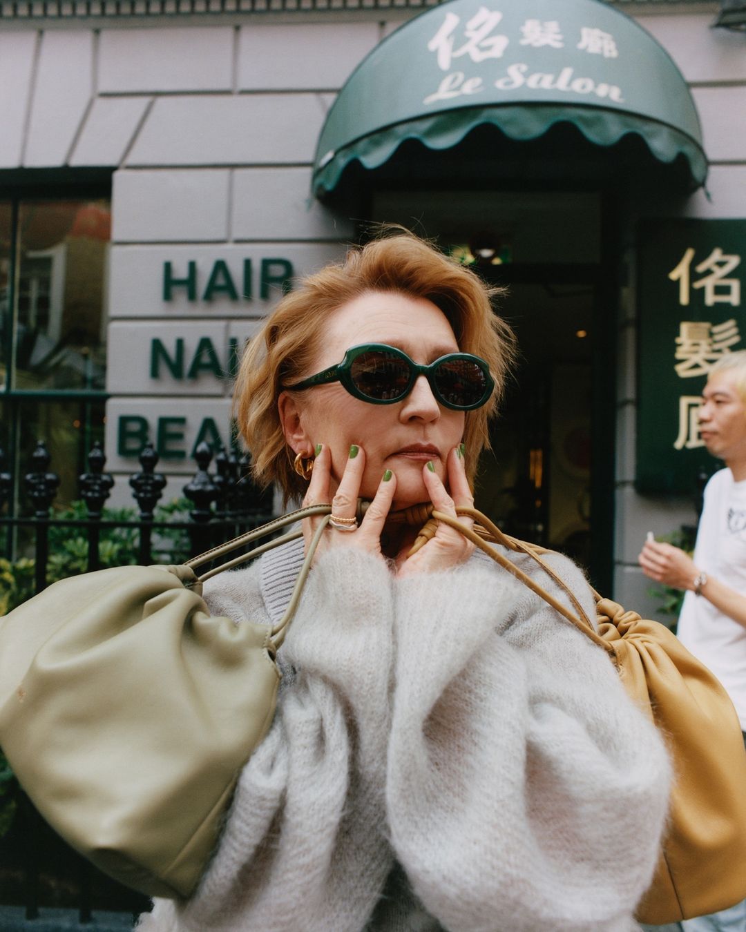 Woman with reddish-blonde hair, wearing a grey sweater and sunglasses, stands in front of a hair salon with two handbags. 