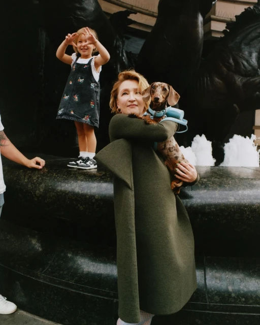 Woman in green coat holds dachshund near child in dress by a fountain; street scene with people, city buildings, and water.