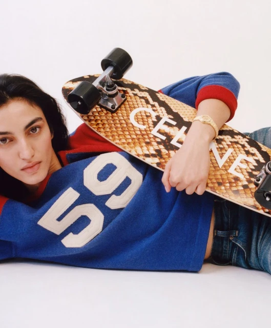 Young woman lying down wearing a blue and red sweater with a snakeskin-patterned skateboard in a studio with a white backdrop.