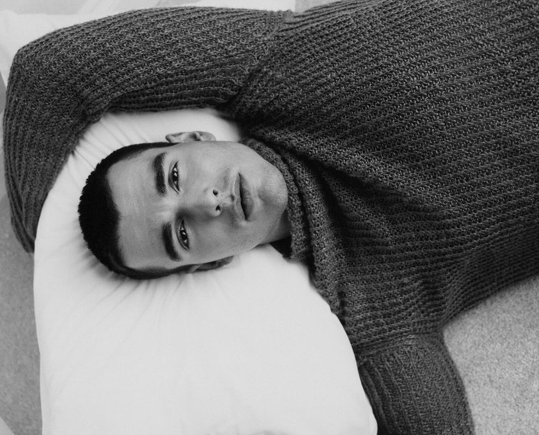 Portrait of a young adult man in a grey knit sweater lying on a white pillow, showcasing a fashion editorial style and a focus on portraiture.