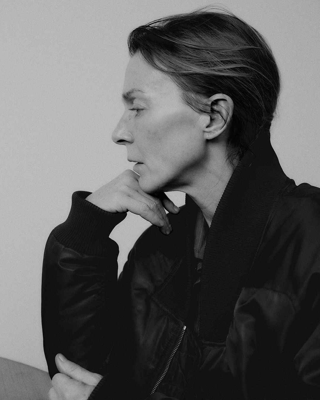 Black and white portrait of an adult woman in profile, wearing a black bomber jacket, resting her chin on her hand against a neutral backdrop.